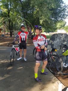 Two cyclists in 'Team NEO' jerseys at a park (rest stop).