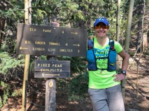 Woman (Coach Nicole) with blue baseball hat, fluorescent green/yellow shirt, gray shorts, and blue hydration vest leaning on a wooden sign. "Pikes Peak Summit 6" "Elk Park 5" "Oil Creek Tunnel 4" Timber line shelter 3"