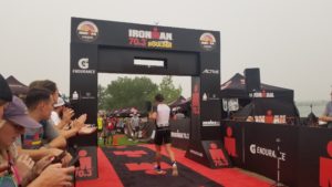 Finish arch of the 2021 Boulder 70.3 race. Arch is black with the race logo. On the ground is red and black carpet with the ironman logo. Spectators are cheering on the finisher going through.