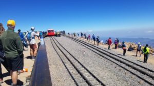 People lined up along cog rail tracks with a red train car in the background. The sky is bright blue with a thin layer of clouds below.