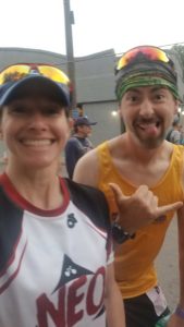 Nicole in front left smiling, wearing blue baseball hat with sunglasses on the brim, slightly behind her on the right of the image is a man in a yellow tank top, green headband sticking out his tongue and giving the hang loose hand sign.