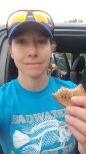 Woman (Nicole) with teal shirt that reads Badwater and a picture of a fish skeleton, hat and sunglasses on the bring. She's holding a section of a sandwich.