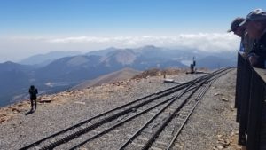 View from the Pikes Peak summit - people looking over a railing at the cog rail tracks, the mountains are below, with clouds in the distance. The sky above is blue!