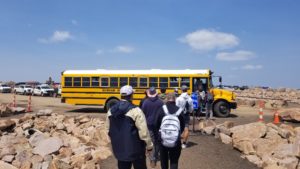 People lined up to get on a yellow school bus at the top of Pikes Peak. Blue-ish (a bit hazy) sky and some small puffy clouds in the background.