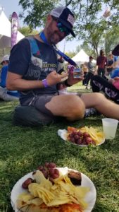 Rudy wearing blue t-shirt and his hydration vest, hat and sunglasses, sitting in the green grass with a plate of food and cup of lemonade. In the foreground is another plate of food (potato chips, pizza, grapes, and a brownie)