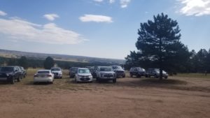 View of a dirt field with nearly a dozen cars/trucks parked. There is a evergreen tree on the right side of the image. The sky is blue with a few clouds.