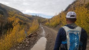 Overcast sky on a rough dirt mountain road with puddles in the ruts. In foreground man with blue and white hydration pack, blue windbreaker, and white trucker hat. Trees/brush on the side of the road are golden, giving the whole photo a brown/gold/yellow feel