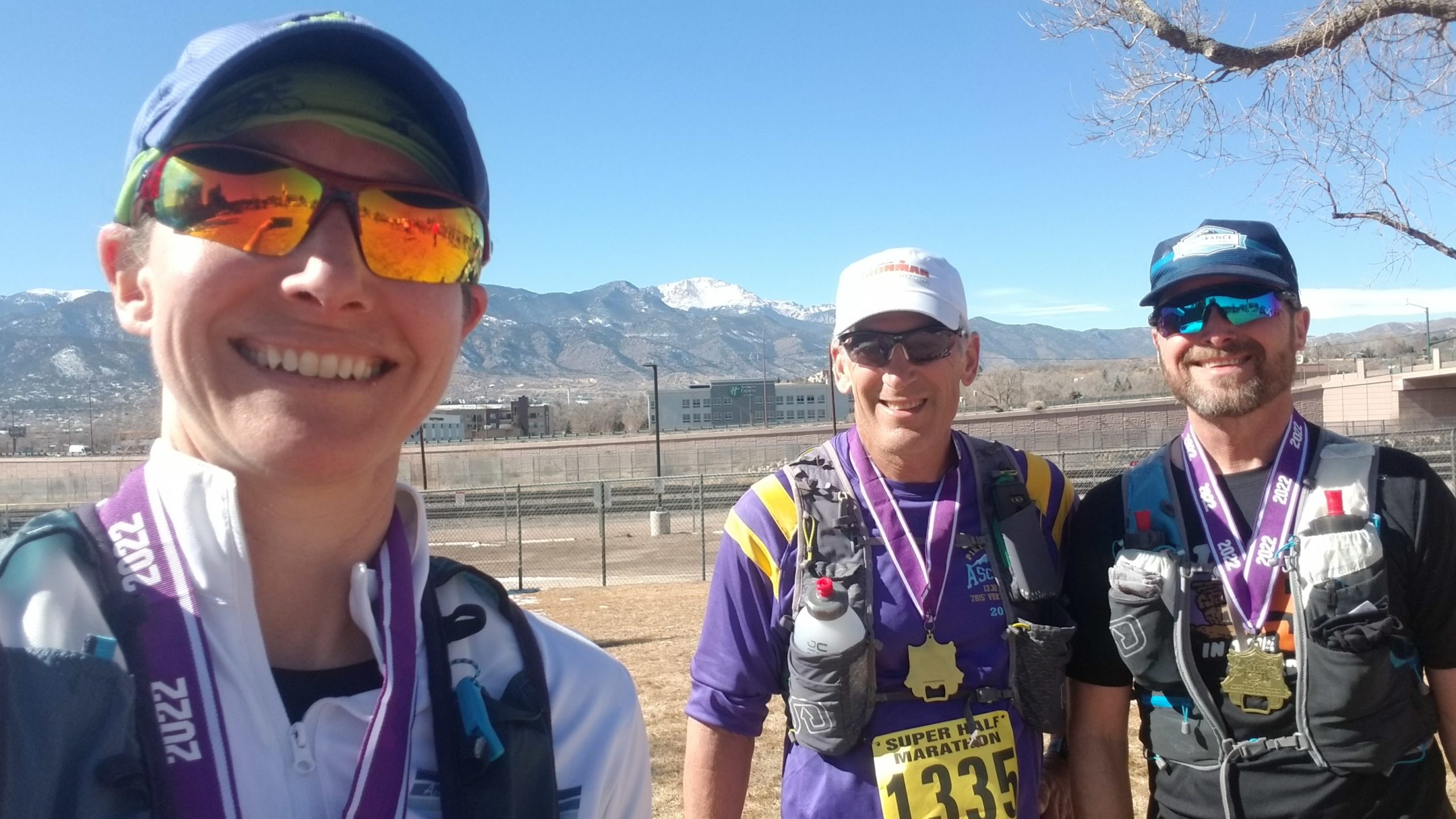 selfie of three runners after a race, one woman on the left (photo taker) and two men on the right. All are wearing baseball hats, sunglasses, and race medals. A snow-capped mountain is in the background far off in the distance.