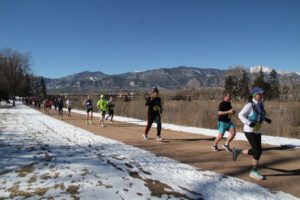 Runners on a flat dirt trail with hills and a snowcapped mountain in the distance. They sky is blue and the ground next to the trail has a thin layer of snow.