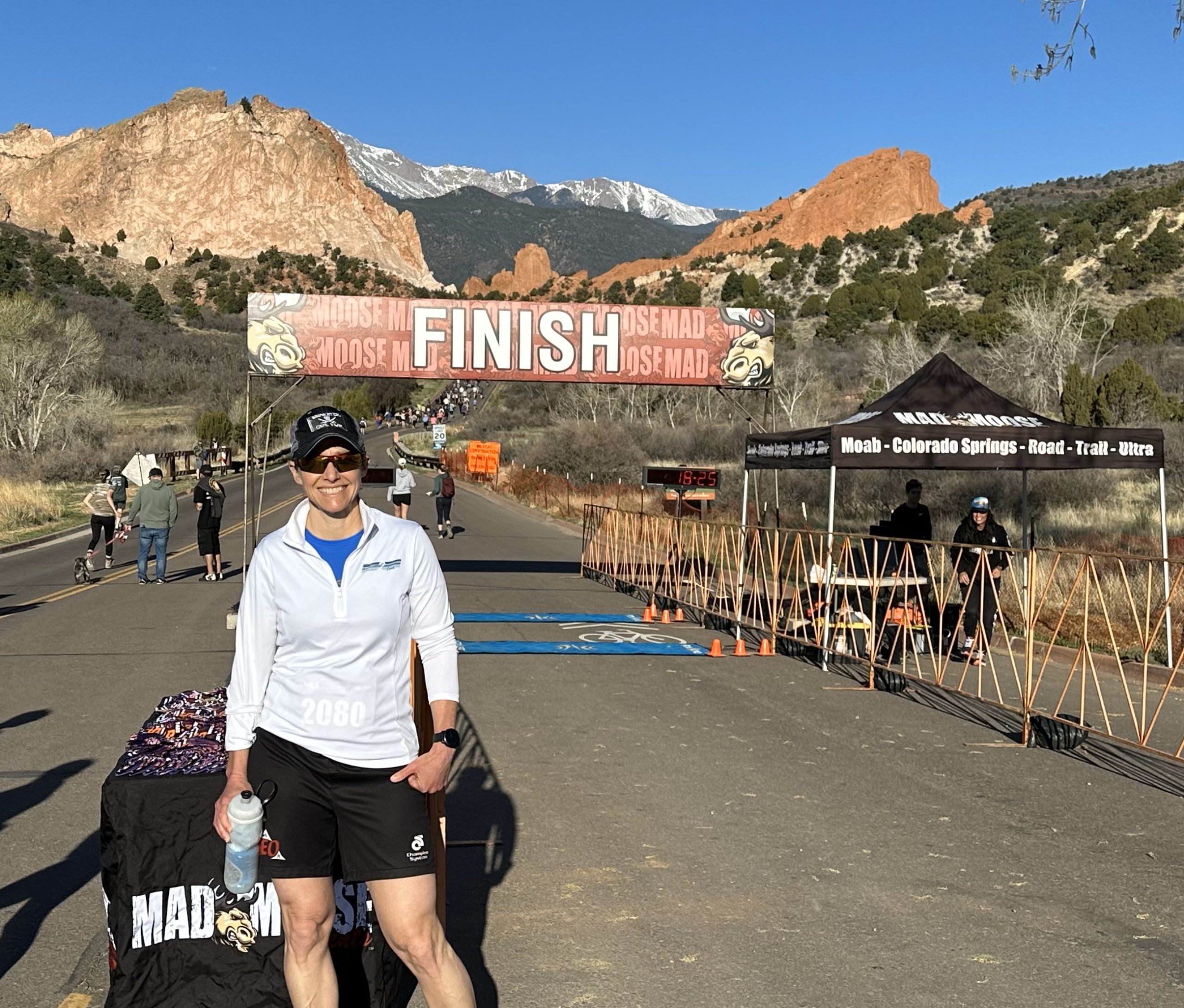 White woman in a white technical pullover, black ball cap, black shorts, and sunglasses is holding an insulated bike water bottle. She’s standing on a road at a race start/finish with the Finish line banner behind her. Further in the distance is mountain scrub, red rocks, and a snow capped mountain.