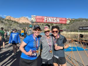 Three white women with sunglasses wearing race medals in front of a finish line banner. Blue skies and red rocks in scrub foothills in the distance.