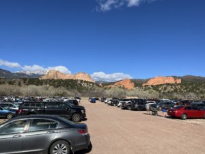 View of red rocks and foothills from a red dirt parking lot filled with cars. A blue sky with some puffy clouds are int he background