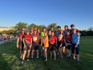 group of 15 people in colorful cycling clothes posing for a group picture on a grass lawn shortly after sunrise.