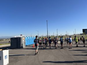 a row of portable toilets in an asphalt parking lot with people waiting in line. The sky is bright blue behind them.