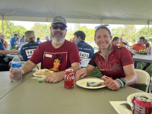 A white man with a moderately long mostly-gray beard, sunglasses, basball had, and red t-shirt is sitting at a lunch table under an outdoor event tent. He's next to a white woman with a red cycling jersey and sunglasses propped on top of her head.