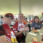 IMG_0805 Two older white men in red, black, and white cycling jerseys and a white woman in a gray t-shirt at a lunch table under an outdoor event tent.