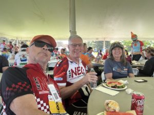 Two older white men in red, black, and white cycling jerseys and a white woman in a gray t-shirt at a lunch table under an outdoor event tent.