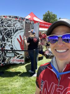 selfie of a white woman in a baseball hat, sunglasses, and red cycling jersey with the left hand brewing beer trailer in the background.