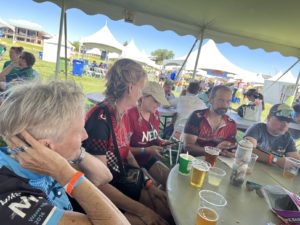 a group of peoplein cycling jerseys sitting at an outdoor table under and event tent with plastic cups filled with beer.