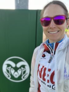 selfie of a white woman with brown hair pulled back, red sunglasses, white and red cycling jersey with a white partially transparent rain jackt. A green box with the Colorado State University ram logo painted in white is in the background