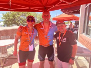 a white female and two white males in cycling kits posing for the camera. They are under an orange outdoor popup tent so there is an orange hue to the image.