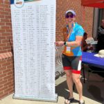IMG_6290 white woman in blue cap, sunglasses, cycling kit, and flip flops pointing to a name on a sign with the 2022 Top 150 Club names