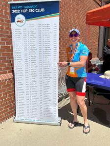 white woman in blue cap, sunglasses, cycling kit, and flip flops pointing to a name on a sign with the 2022 Top 150 Club names