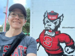 selfie photo of a white woman with glasses and a baseball hat. She's wearing a gray t-shirt and the letters "STATE" are visible in red, with a bag strap across her chest. She's next to the NC State "Tuffy" mascot painted on a white brick building.