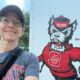 selfie photo of a white woman with glasses and a baseball hat. She's wearing a gray t-shirt and the letters "STATE" are visible in red, with a bag strap across her chest. She's next to the NC State "Tuffy" mascot painted on a white brick building.