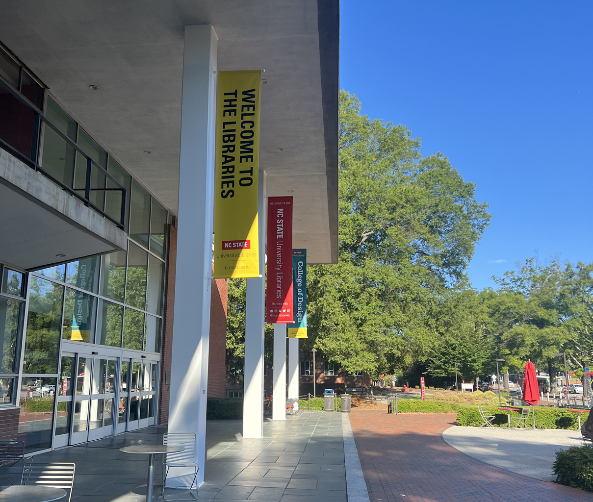 Exterior of a library with glass windows, pillars, and a stone plaza entry adjacent to a brick walkway.