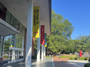 Exterior of a library with glass windows, pillars, and a stone plaza entry adjacent to a brick walkway.